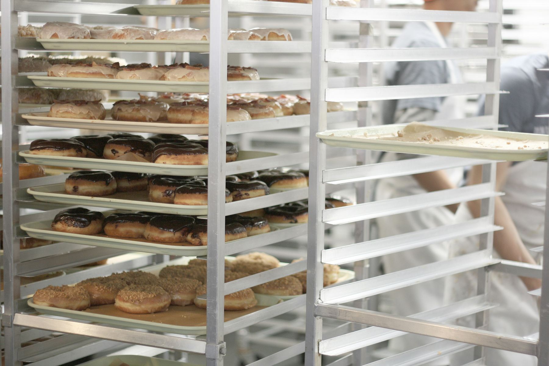 Racks of glazed doughnuts on trays in a commercial bakery, with a baker in whites behind them
